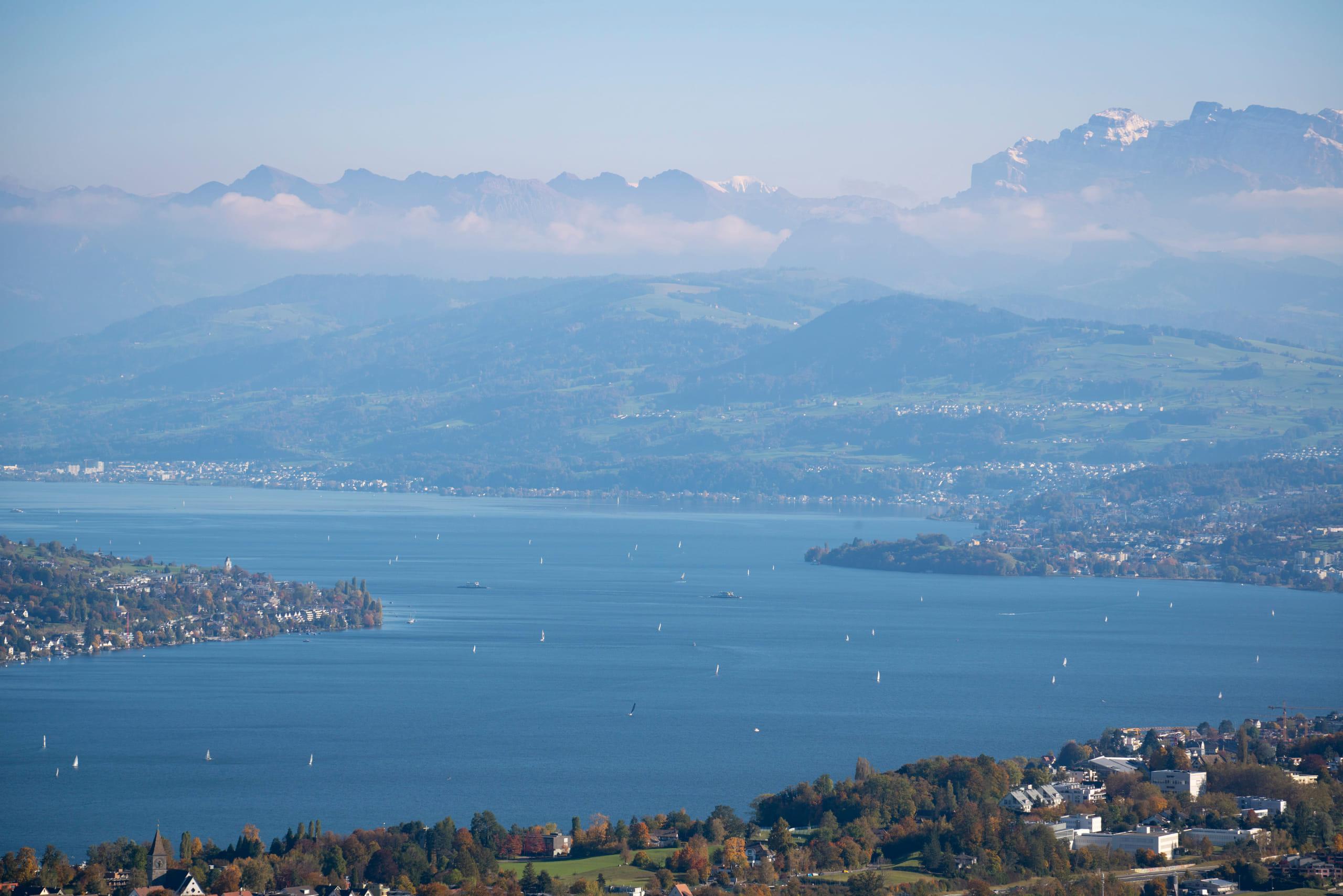 A Swiss lake from above with sailboats scattered across the water and the mountains layered behind