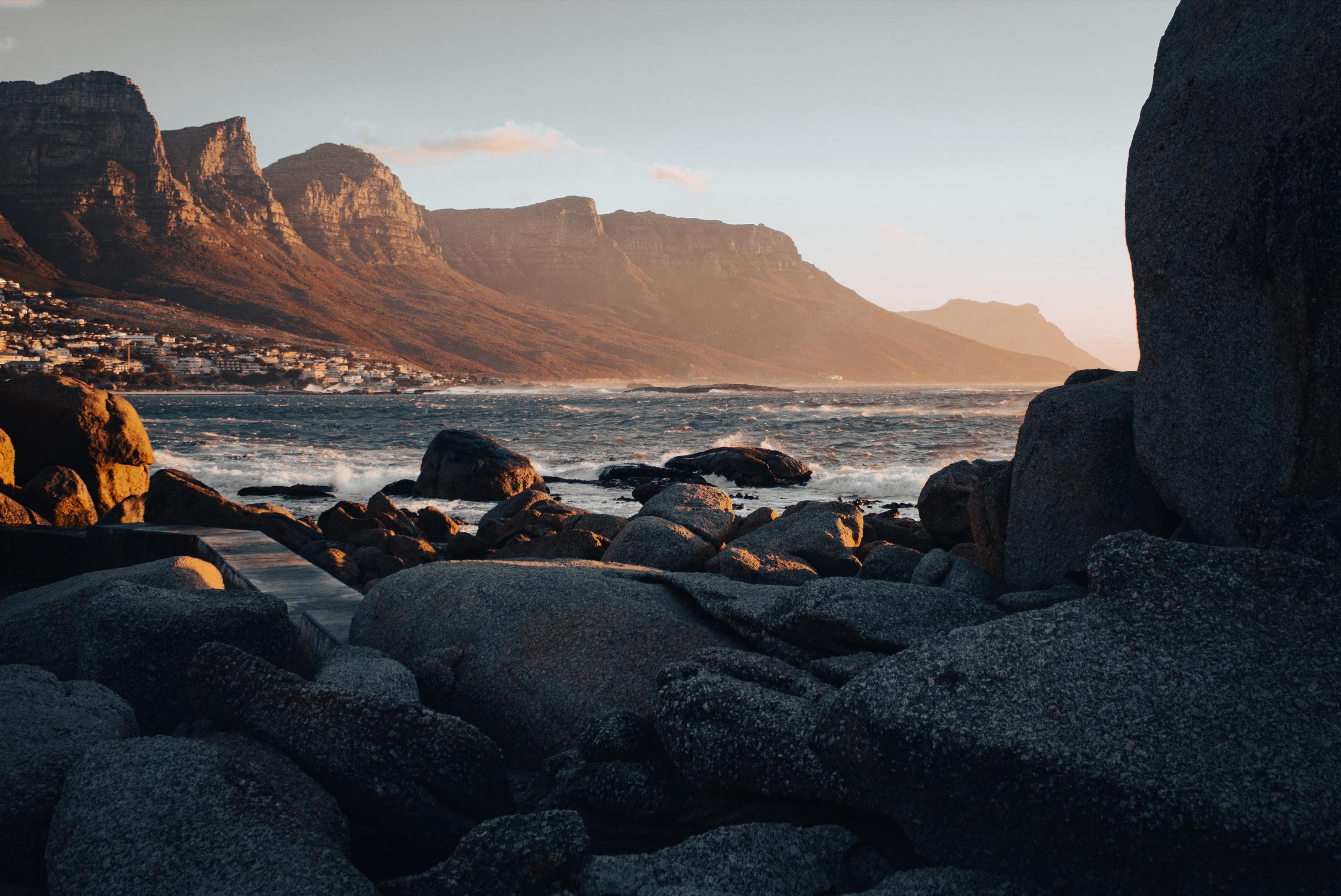 Golden-hour light on the Twelve Apostles mountains above Camps Bay, Cape Town, seen from shadowed granite boulders on the shore