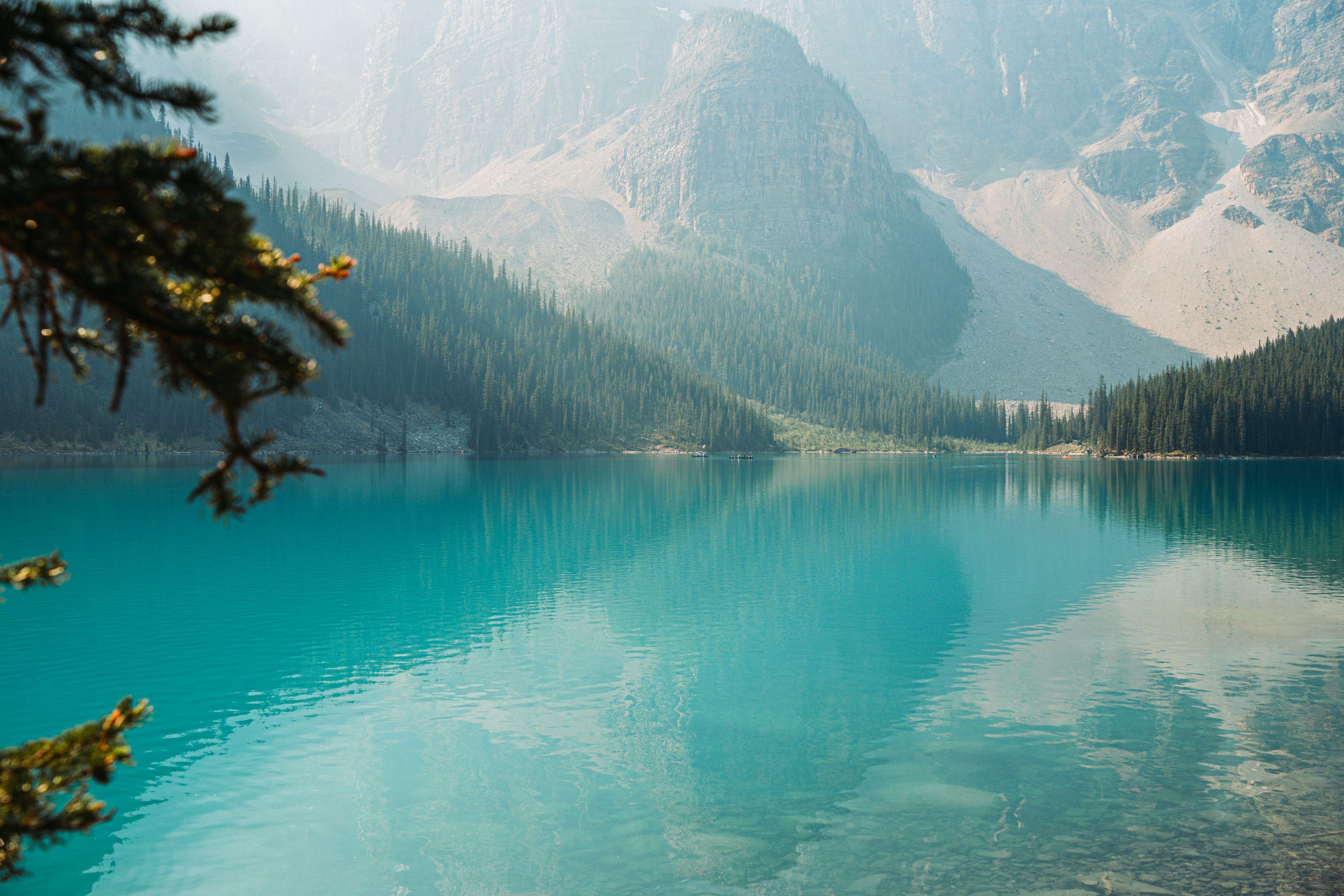A still turquoise mountain lake below hazy grey peaks, dark pines along the far shore