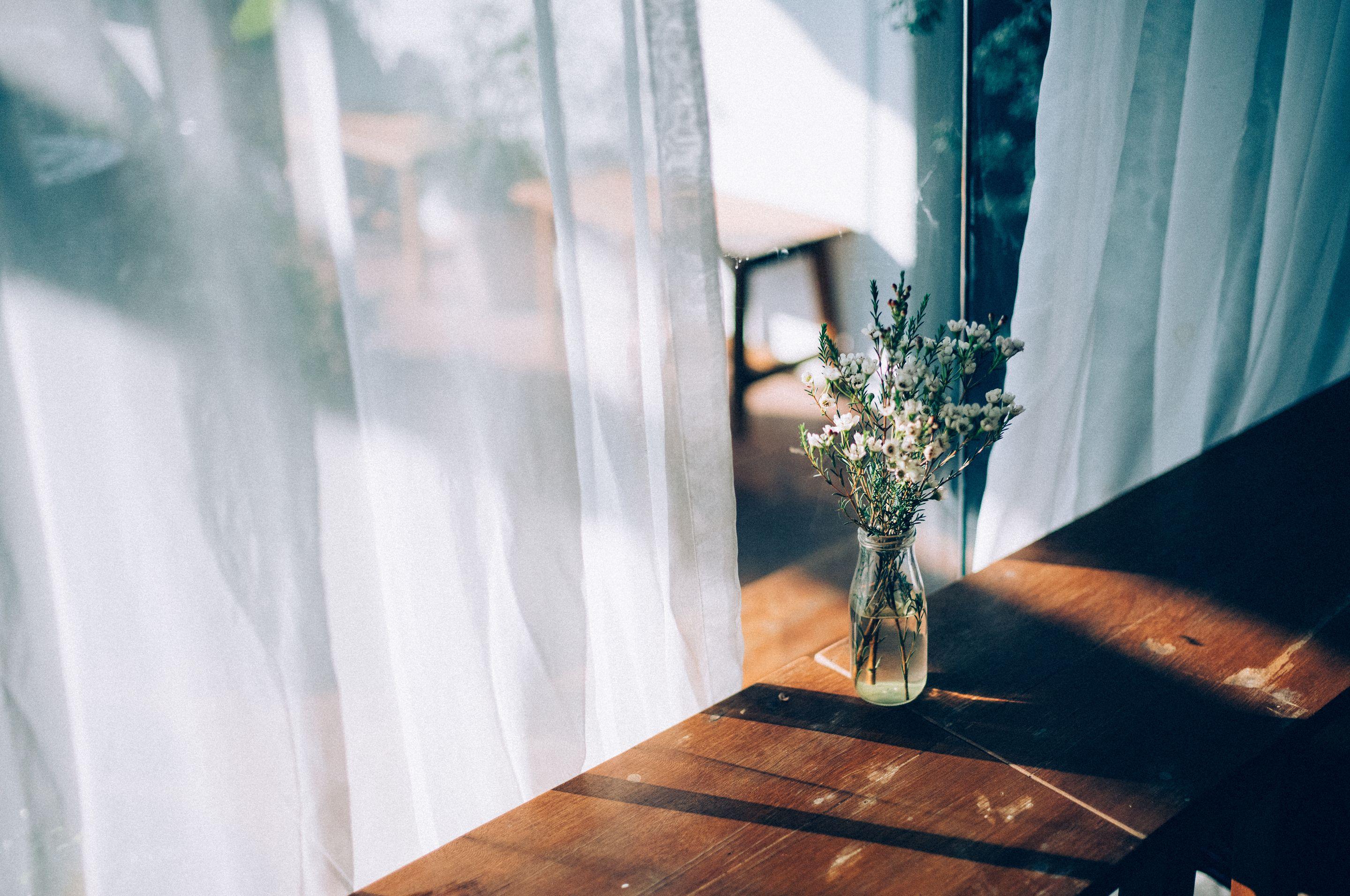 Sunlight through a sheer white curtain onto a wooden table with a jar of dried flowers