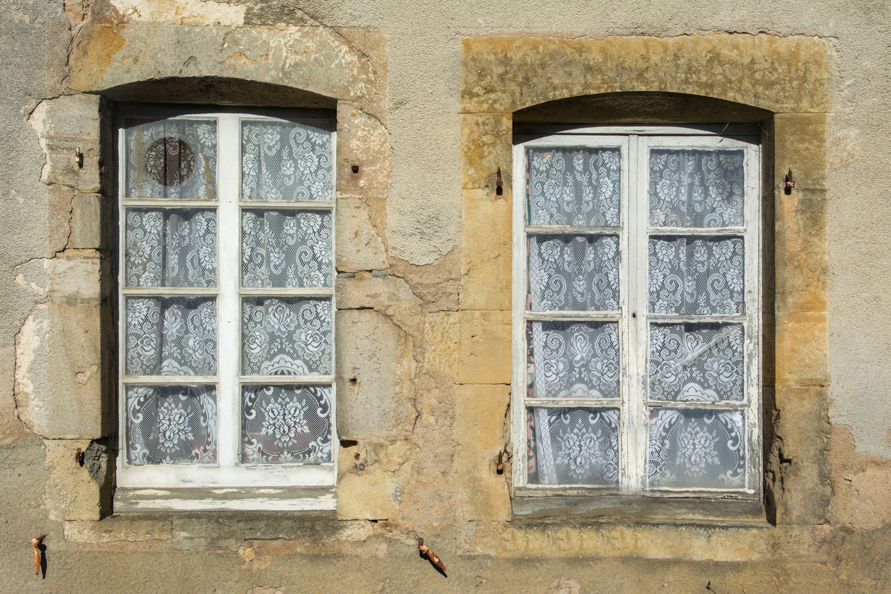 Two lace-curtained windows set in a warm limestone wall in bright daylight