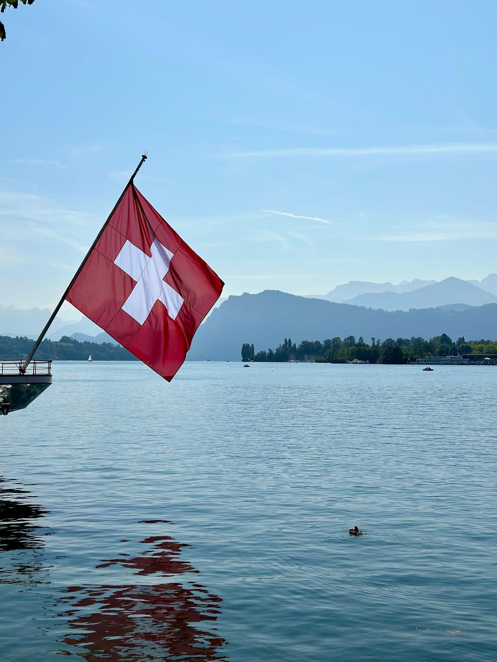 A Swiss flag on the shore of Lake Lucerne under a clear sky, the mountains beyond