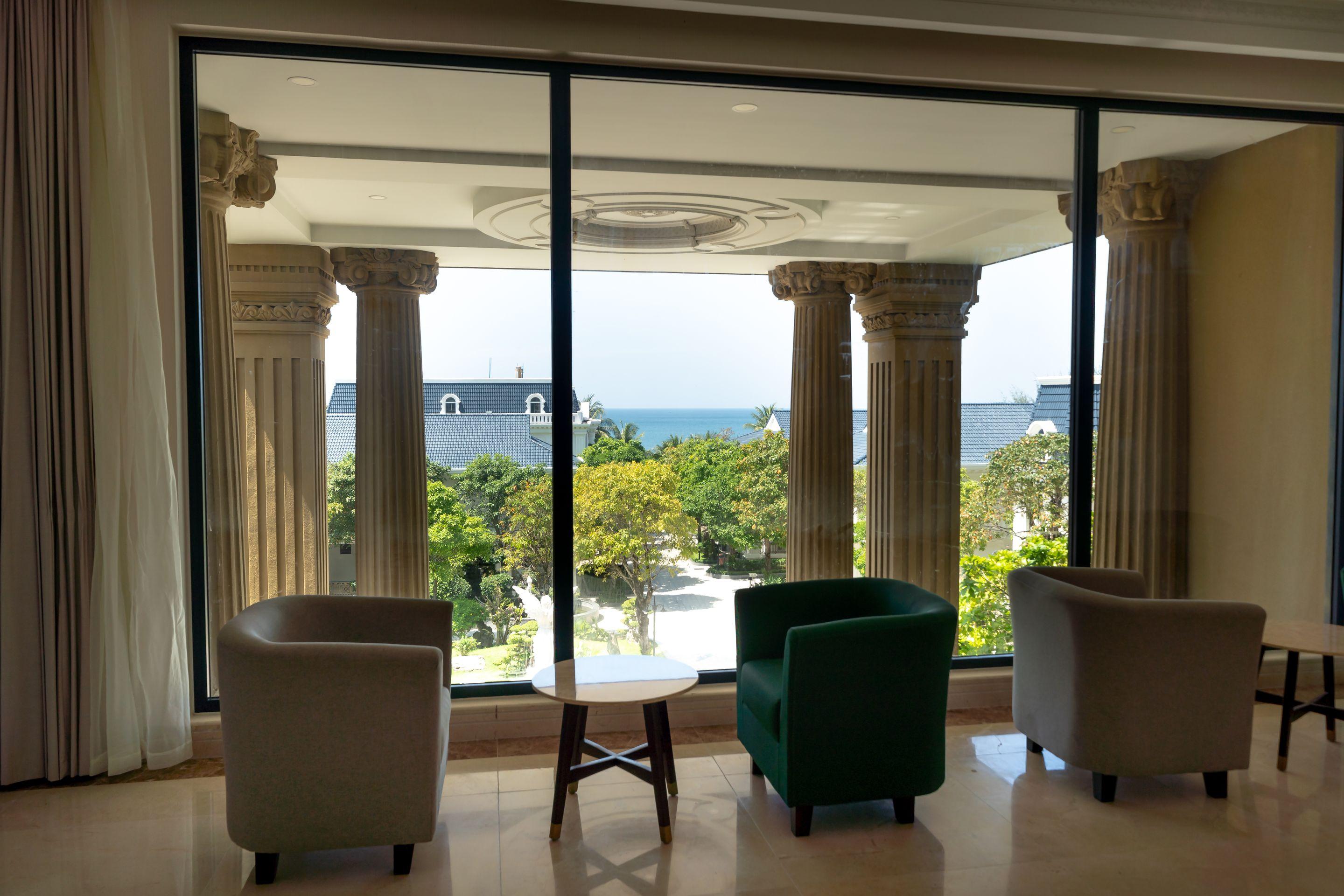 Armchairs beside tall windows in a colonnaded lounge, trees and the sea outside
