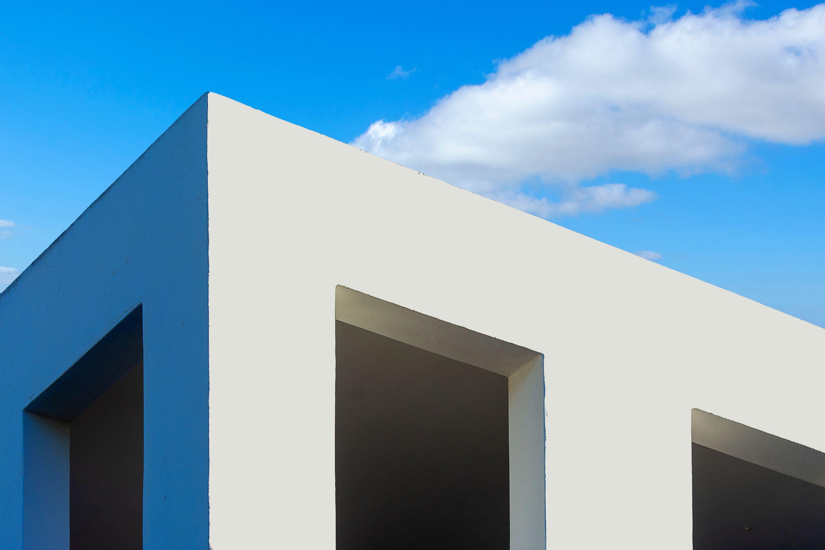 A white cubic building with deep square openings against a blue sky and one small cloud