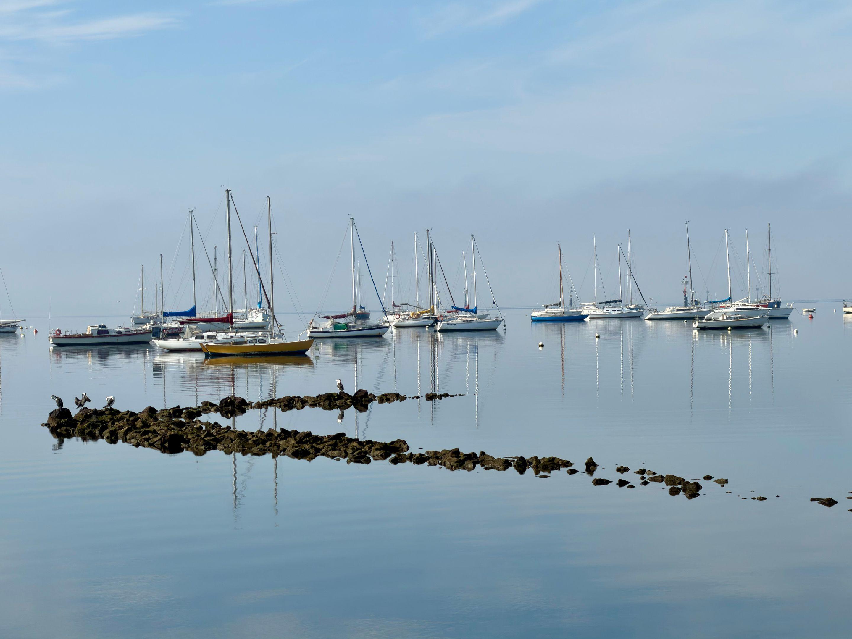 Moored sailing boats on a still pale-blue bay under a soft sky