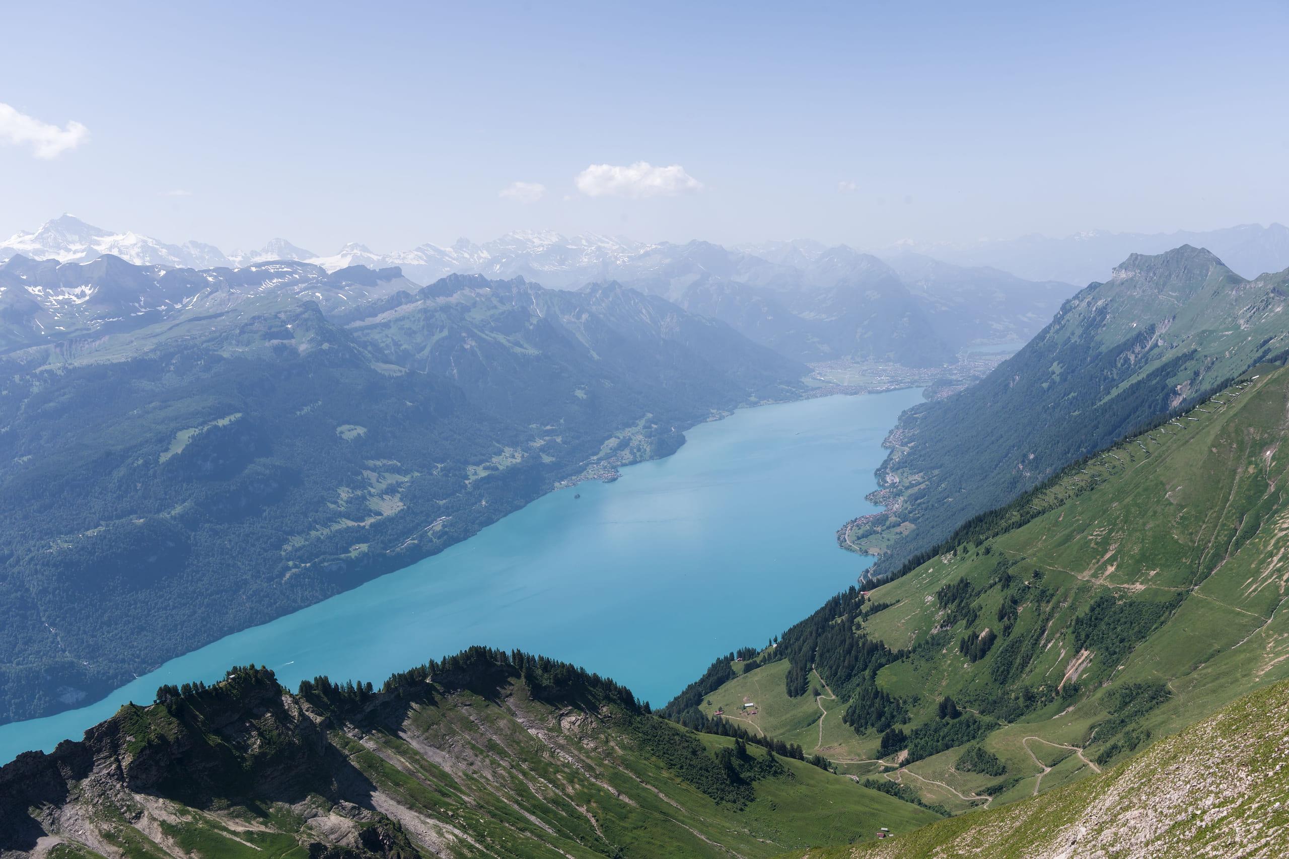 A long turquoise Swiss lake seen from above, held between forested alpine ridges in summer light