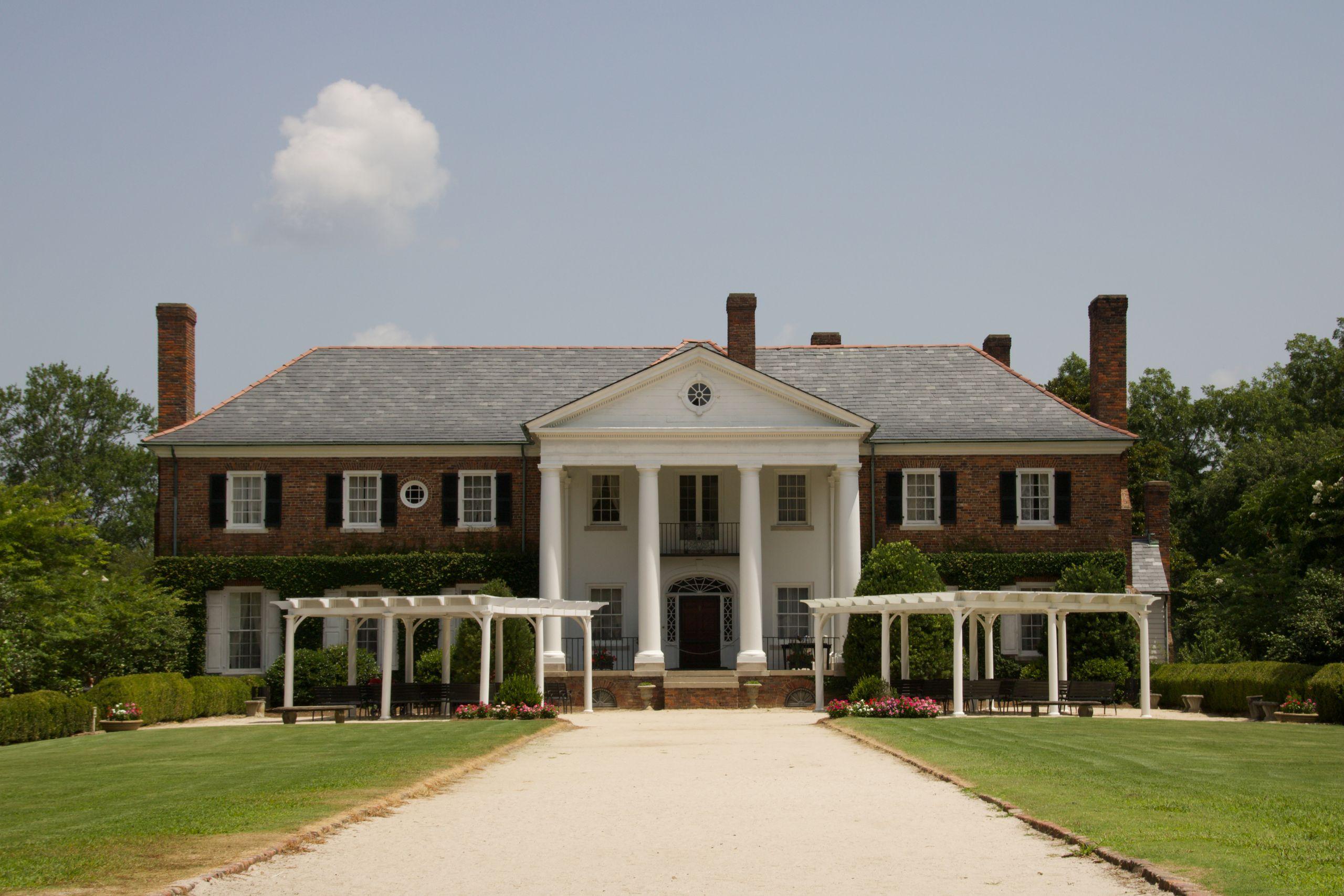 An elegant colonial-style estate with white columns, brick facade, and a manicured lawn under a clear blue sky