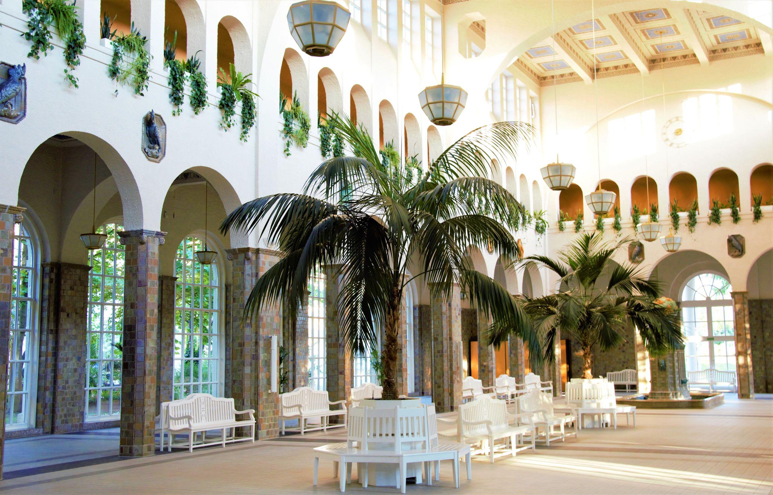 A sunlit atrium with arched colonnades, indoor palm trees, and white benches beneath a vaulted ceiling