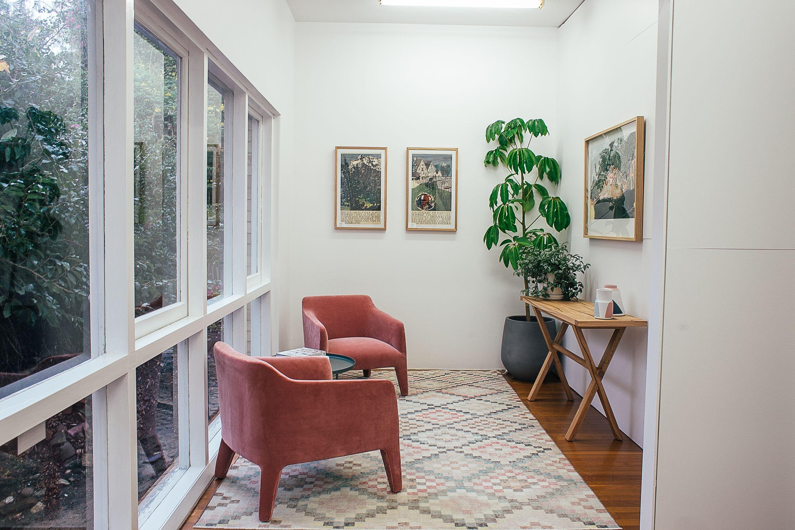 A sunlit room with two soft rose-colored armchairs, a wooden side table, and windows onto a green garden