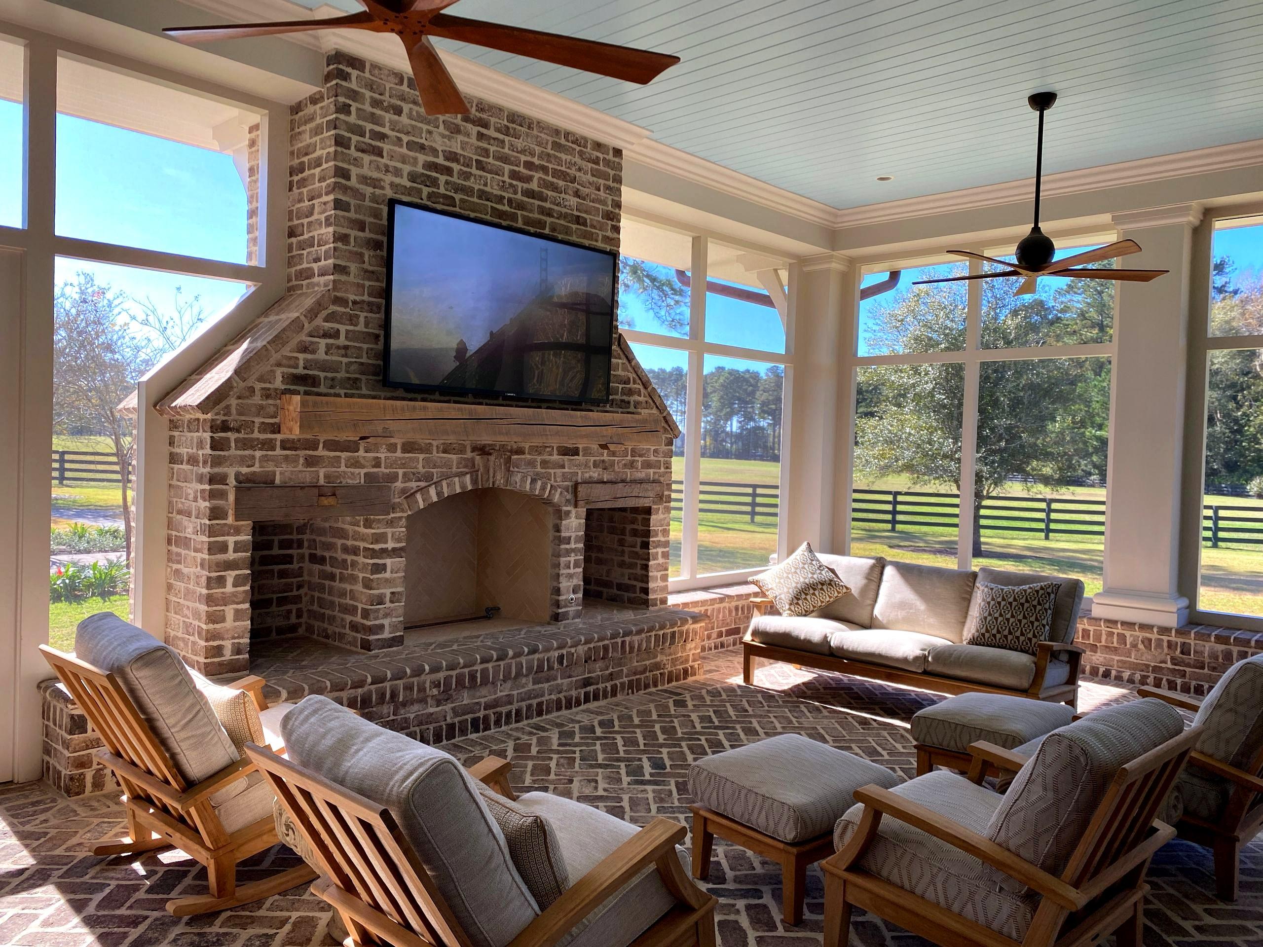 A bright sunroom with walls of windows looking onto green pasture, wooden chairs, and a brick fireplace