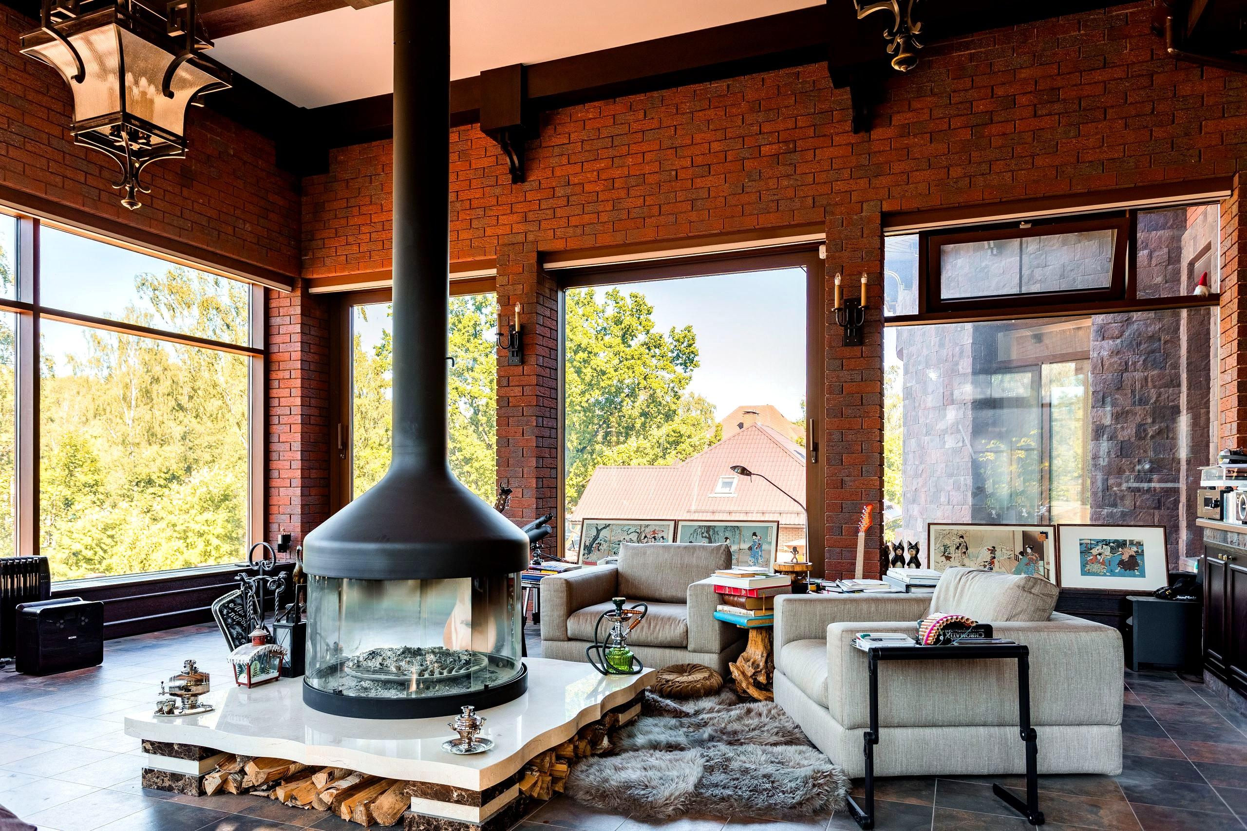 A warm brick-walled living room with a central fireplace, soft sofas, and large windows onto greenery
