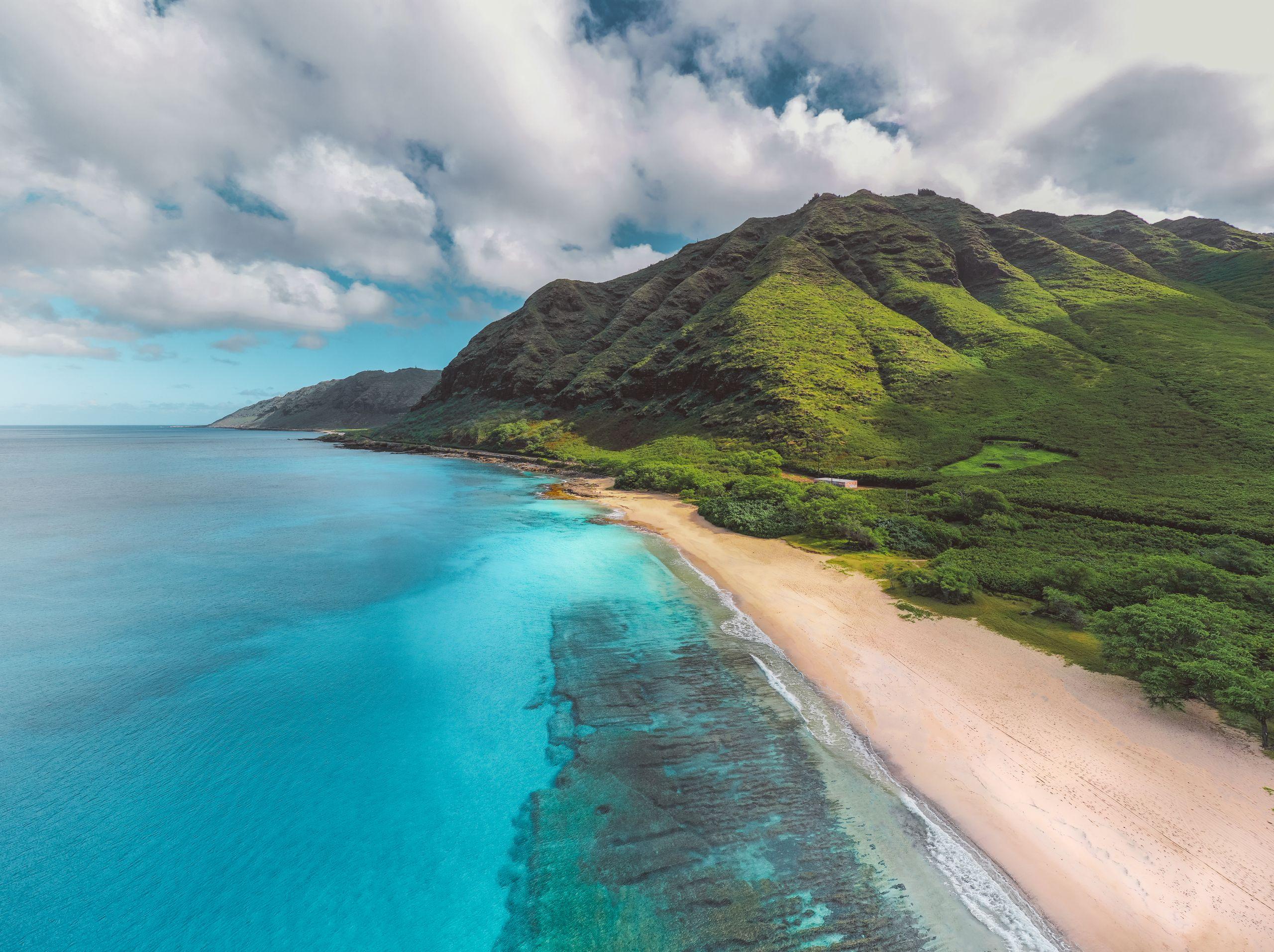 Aerial view of a bright turquoise Hawaiian coastline with a sandy beach and green mountains under a partly clouded sky