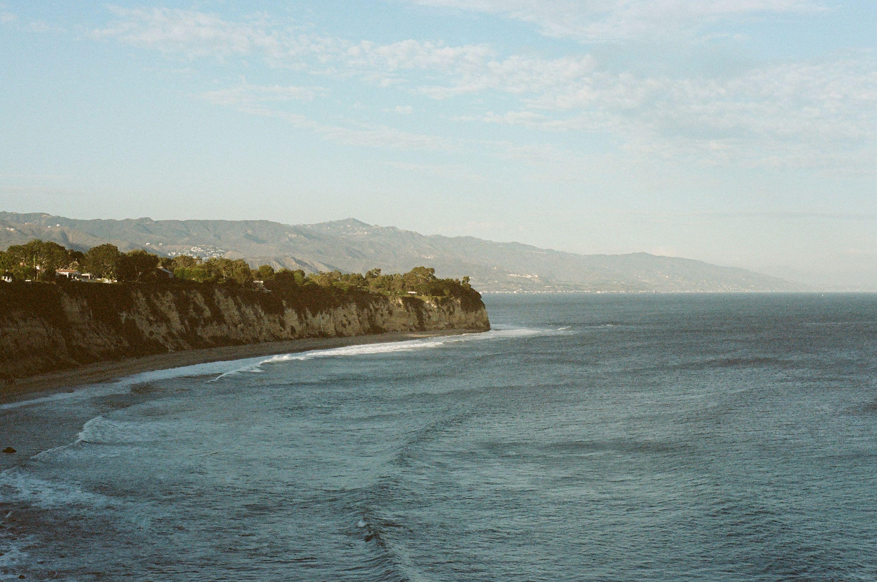 A wooded Malibu headland dropping to a quiet break, the coast range hazy behind