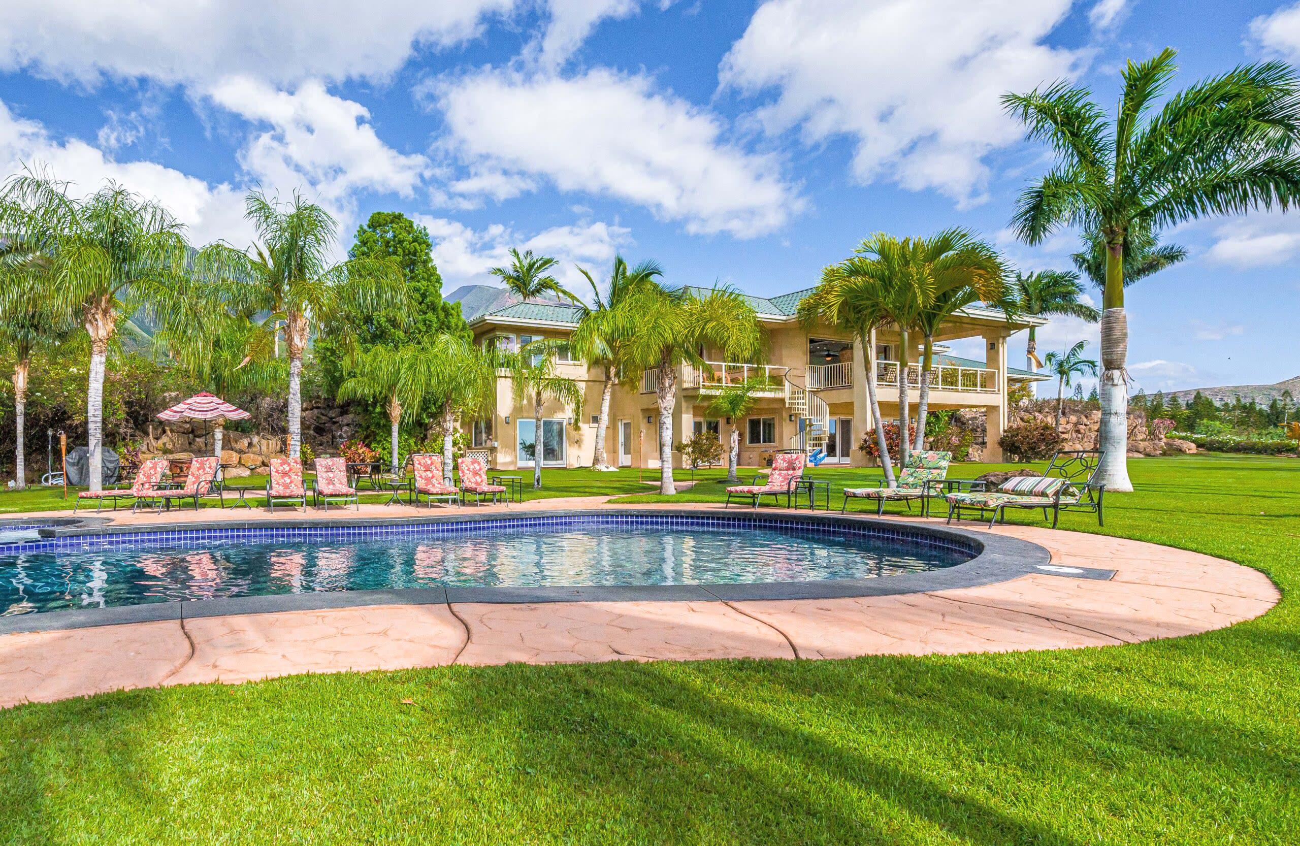 The swimming pool and lawn behind one of Maui Recovery’s homes in Lahaina: lounge chairs, palm trees, and the back of a two-story house with balconies under a blue sky