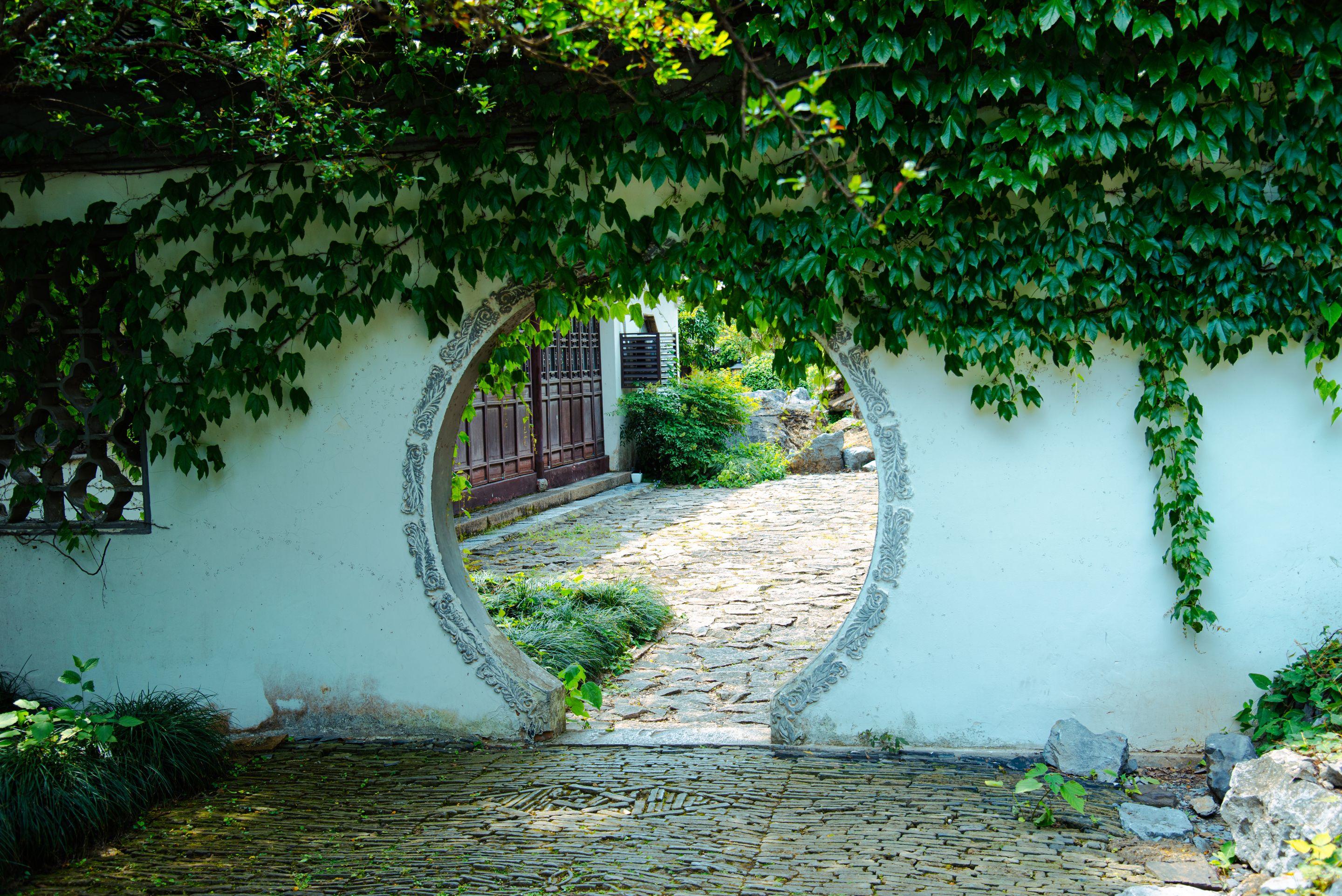 A round moon gate in a pale garden wall opening onto a cobbled path and greenery