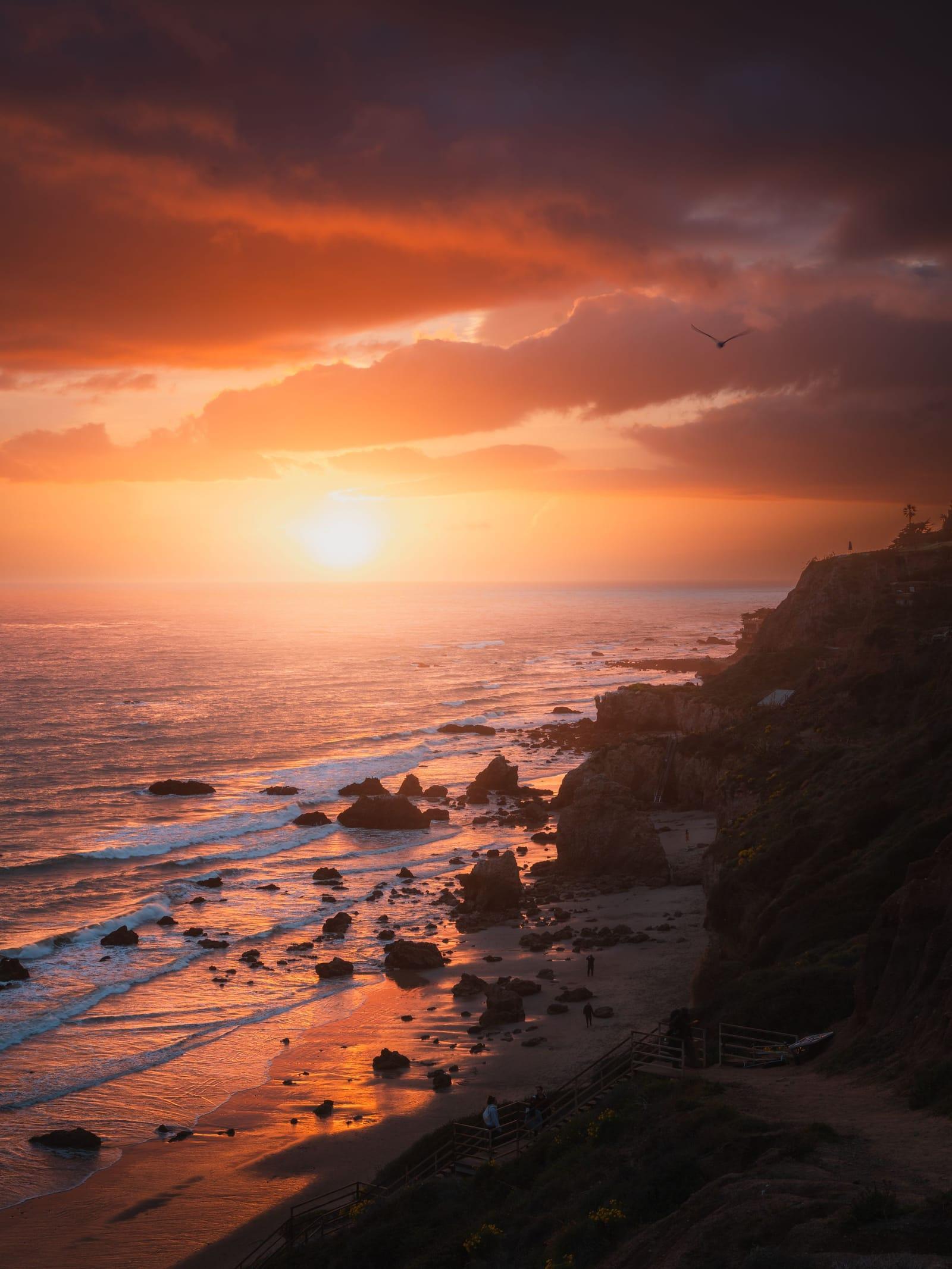 An orange sunset over the rocky Malibu shoreline, waves washing over the rocks