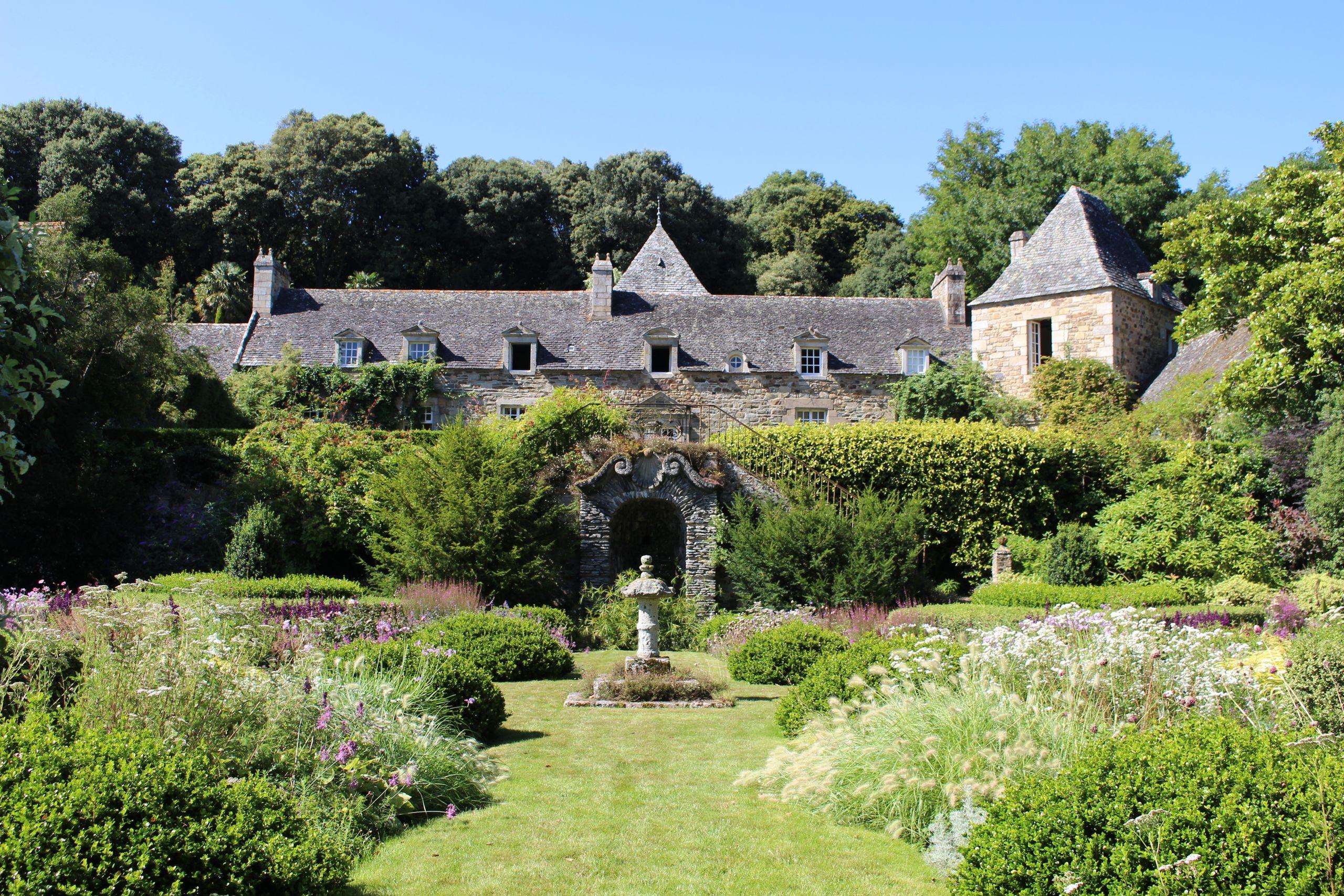 A historic stone manor behind landscaped formal gardens and flowering borders under a clear sky