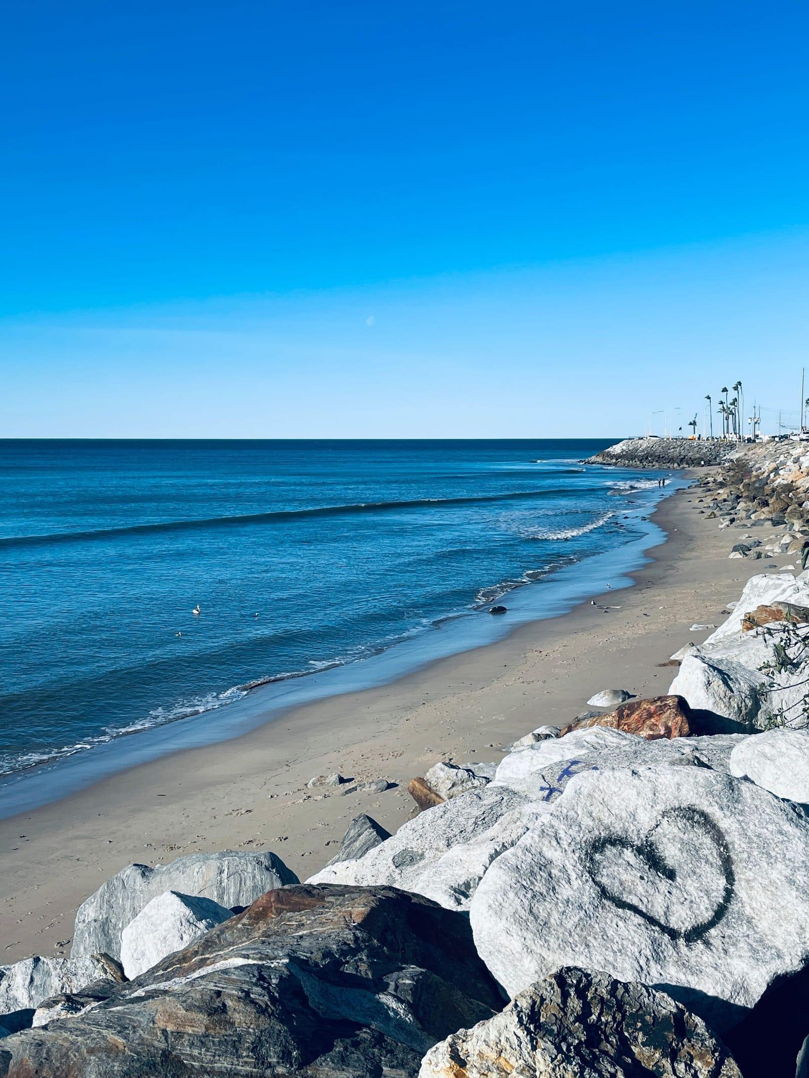 A quiet Malibu beach under a clear blue sky, rocks at the waterline