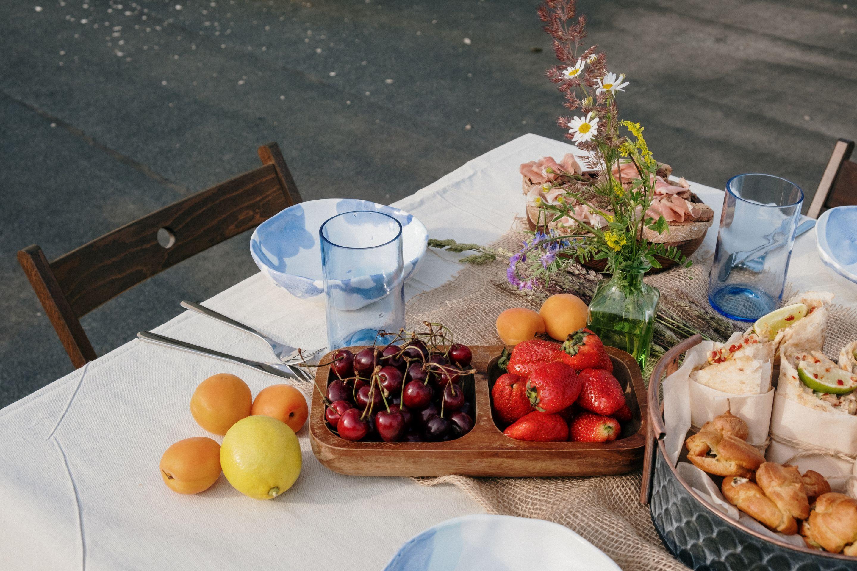 An outdoor table laid with fruit, flowers and glasses on white linen in bright daylight