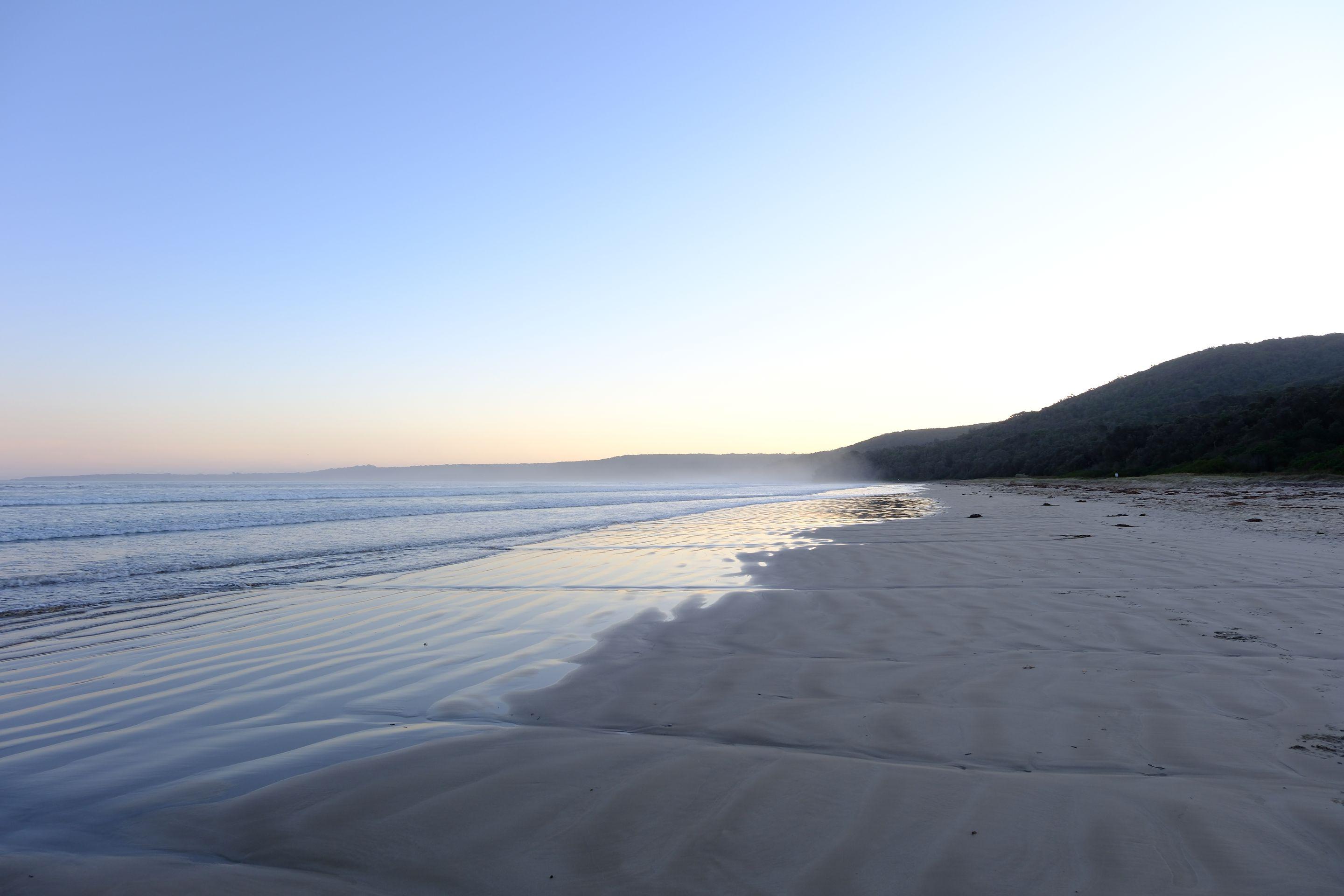 A wide empty beach at first light, pale sky above a calm sea and a low headland