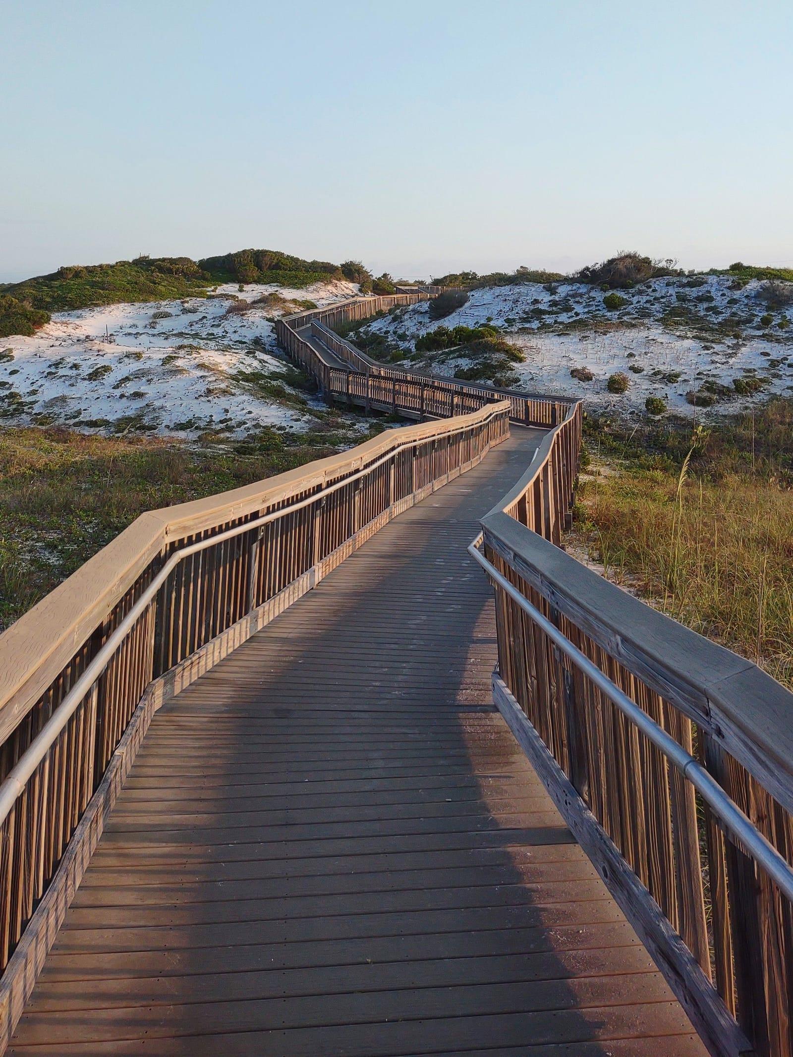 A wooden boardwalk winding over white sand dunes in soft evening light