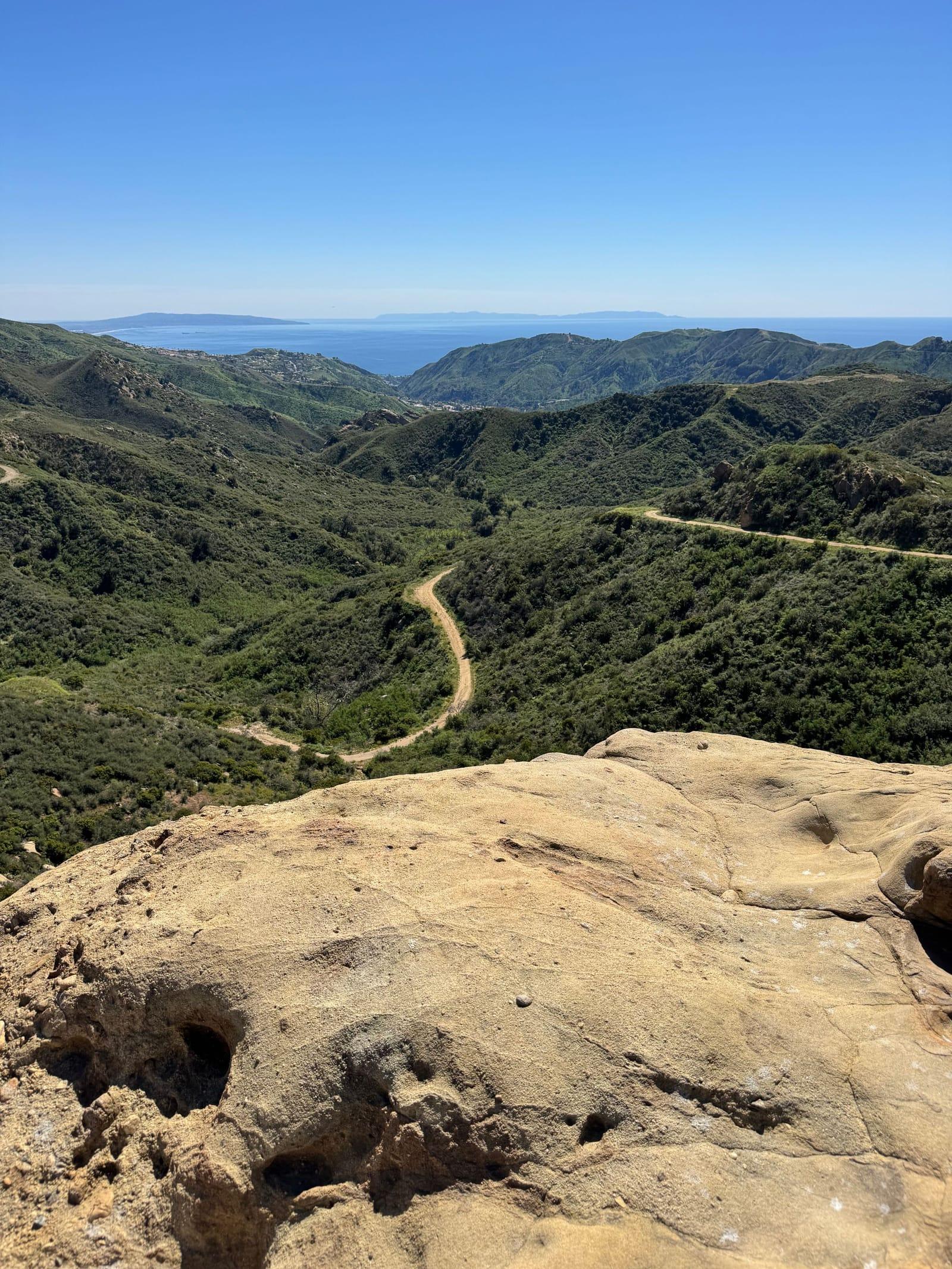 Green canyons of the Santa Monica Mountains running down to the Pacific, a trail winding below a sandstone outcrop