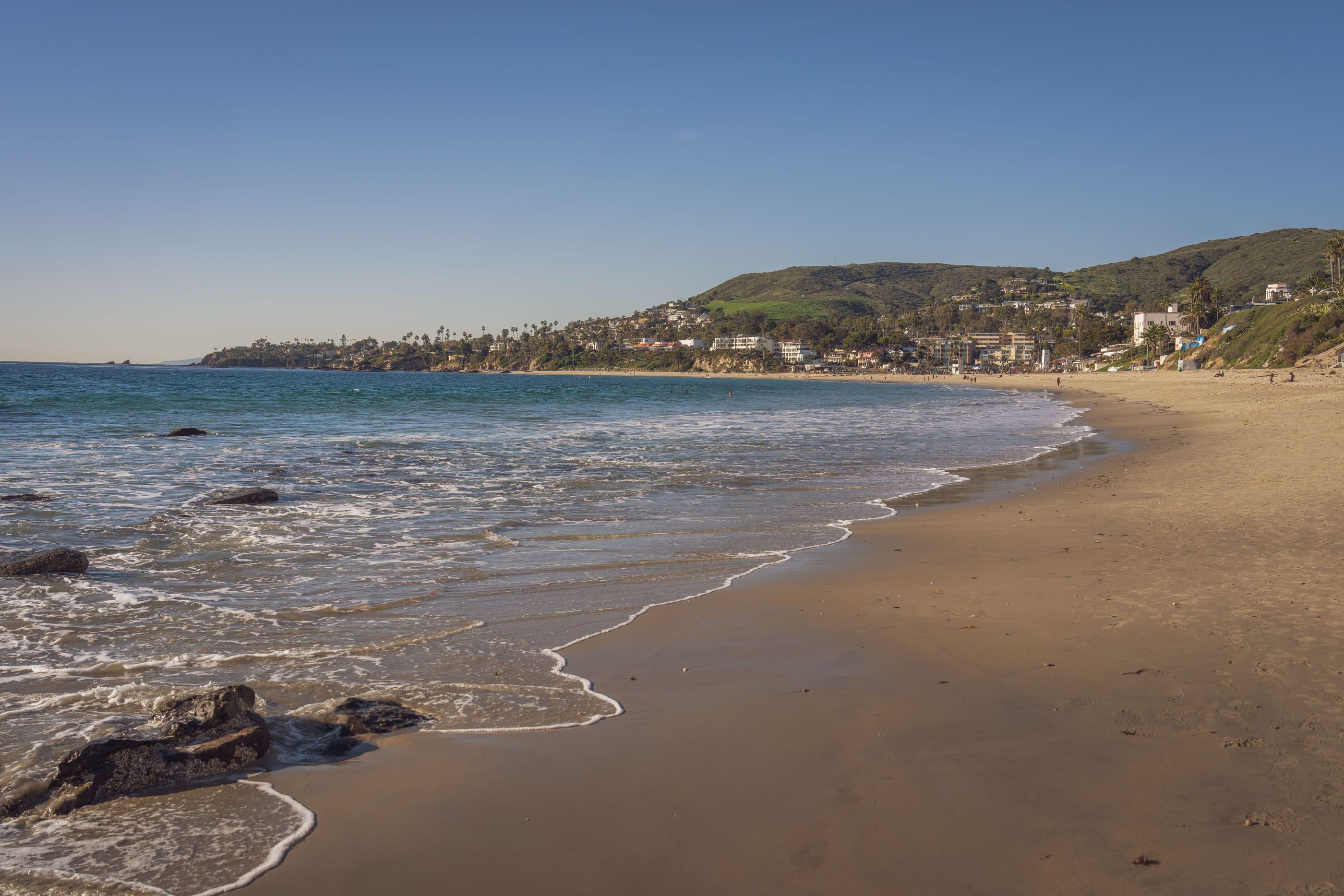 A long California beach below green coastal hills on a clear morning, gentle surf along the sand