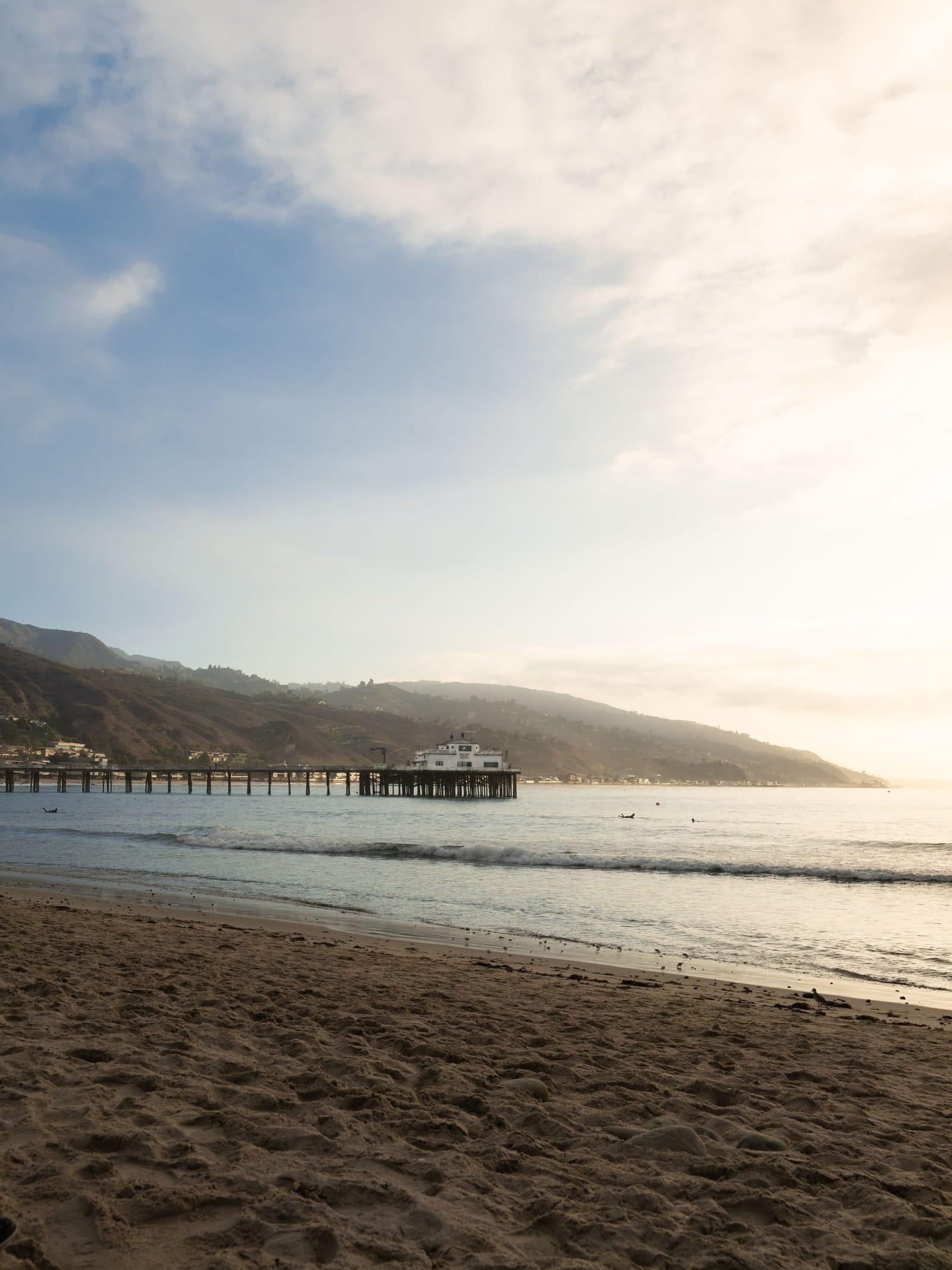 Wet sand and gentle surf on a Malibu beach, the pier and hills beyond in soft evening light