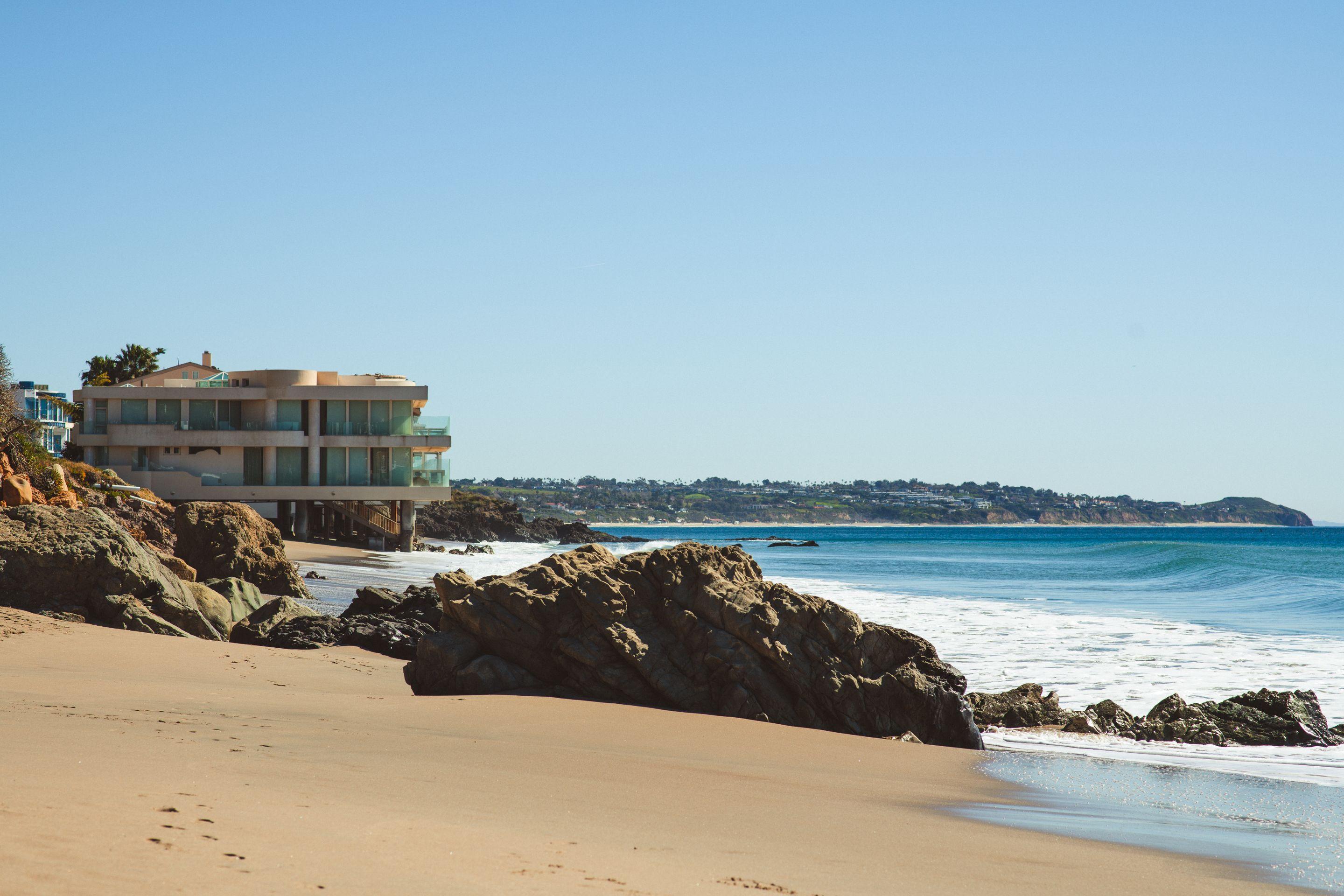A modern house on a Malibu clifftop above an empty beach, ocean stretching to the horizon