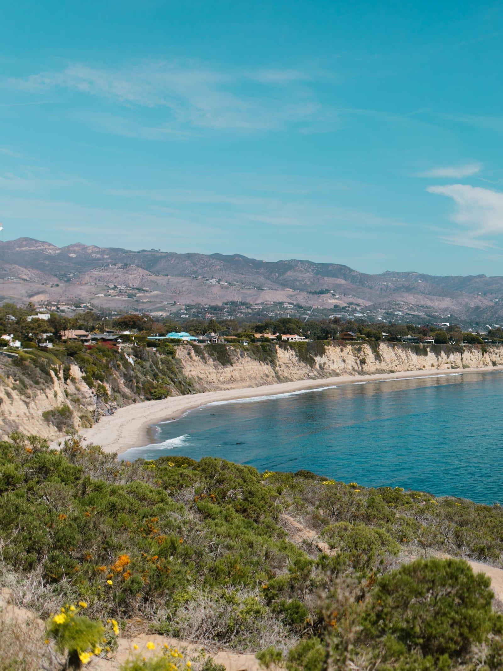 A green bluff above a curving Malibu beach, with mountains in the distance