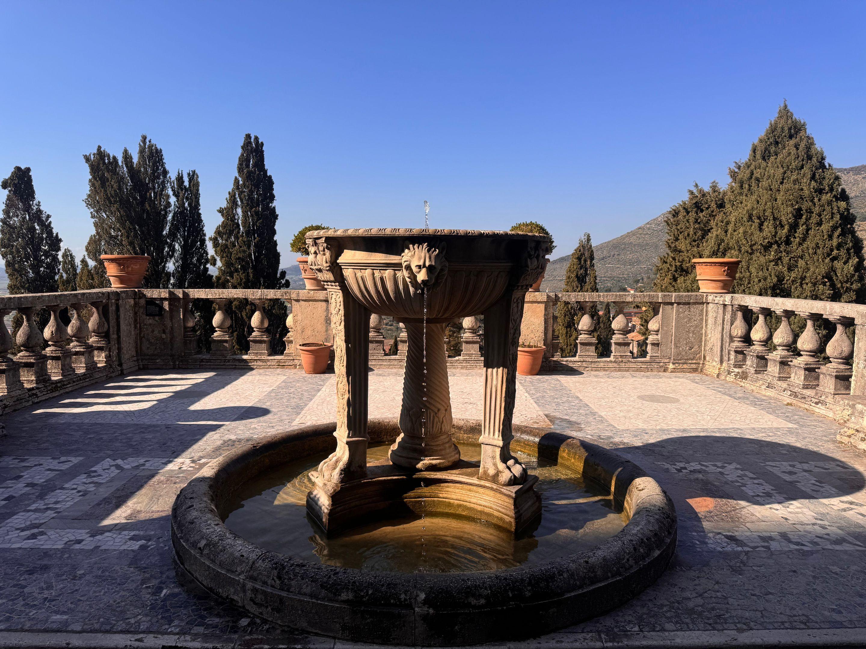 A stone fountain on a wide paved terrace with cypresses and a balustrade behind