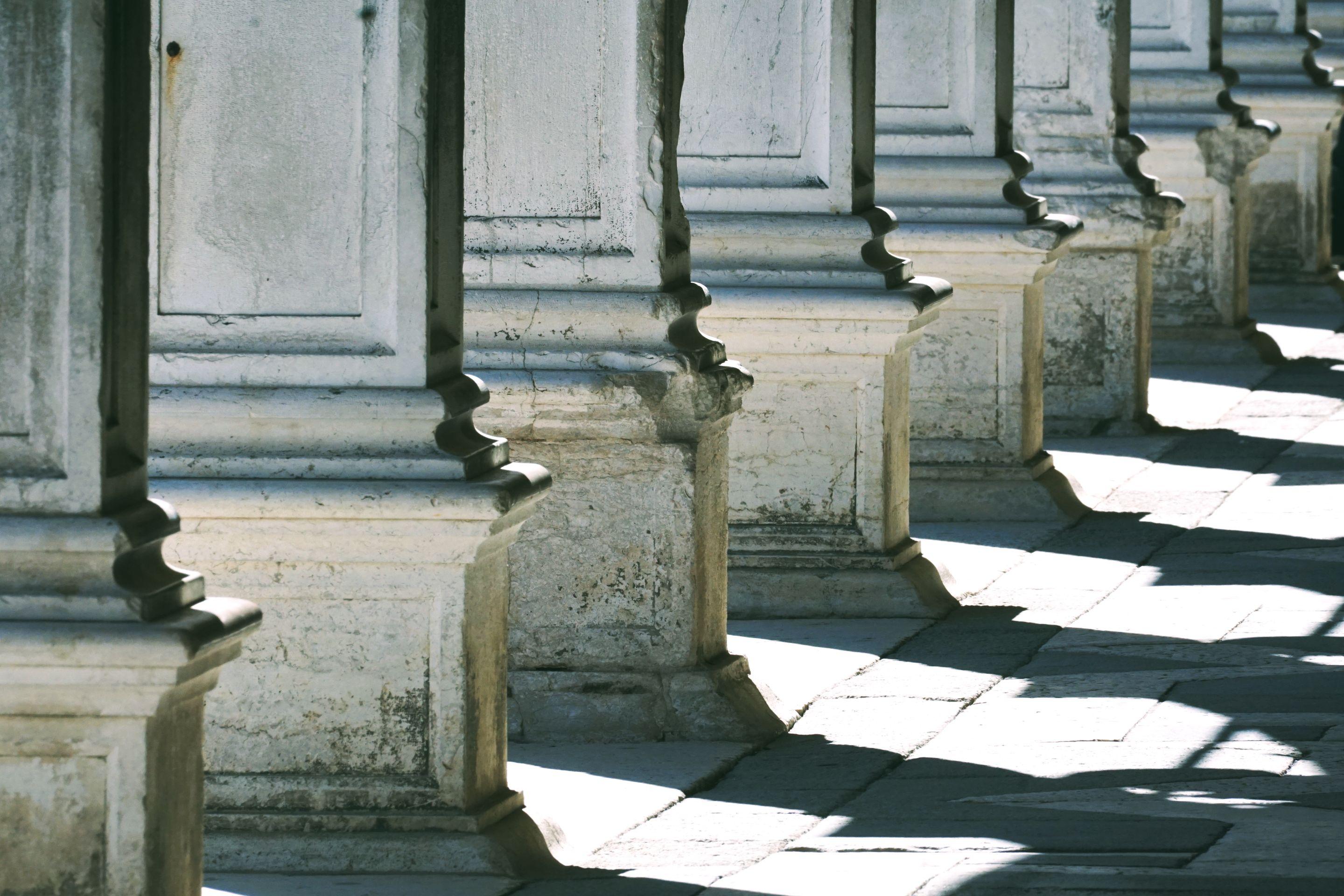 Pale stone columns of an open colonnade, sunlight falling in bands across the paving
