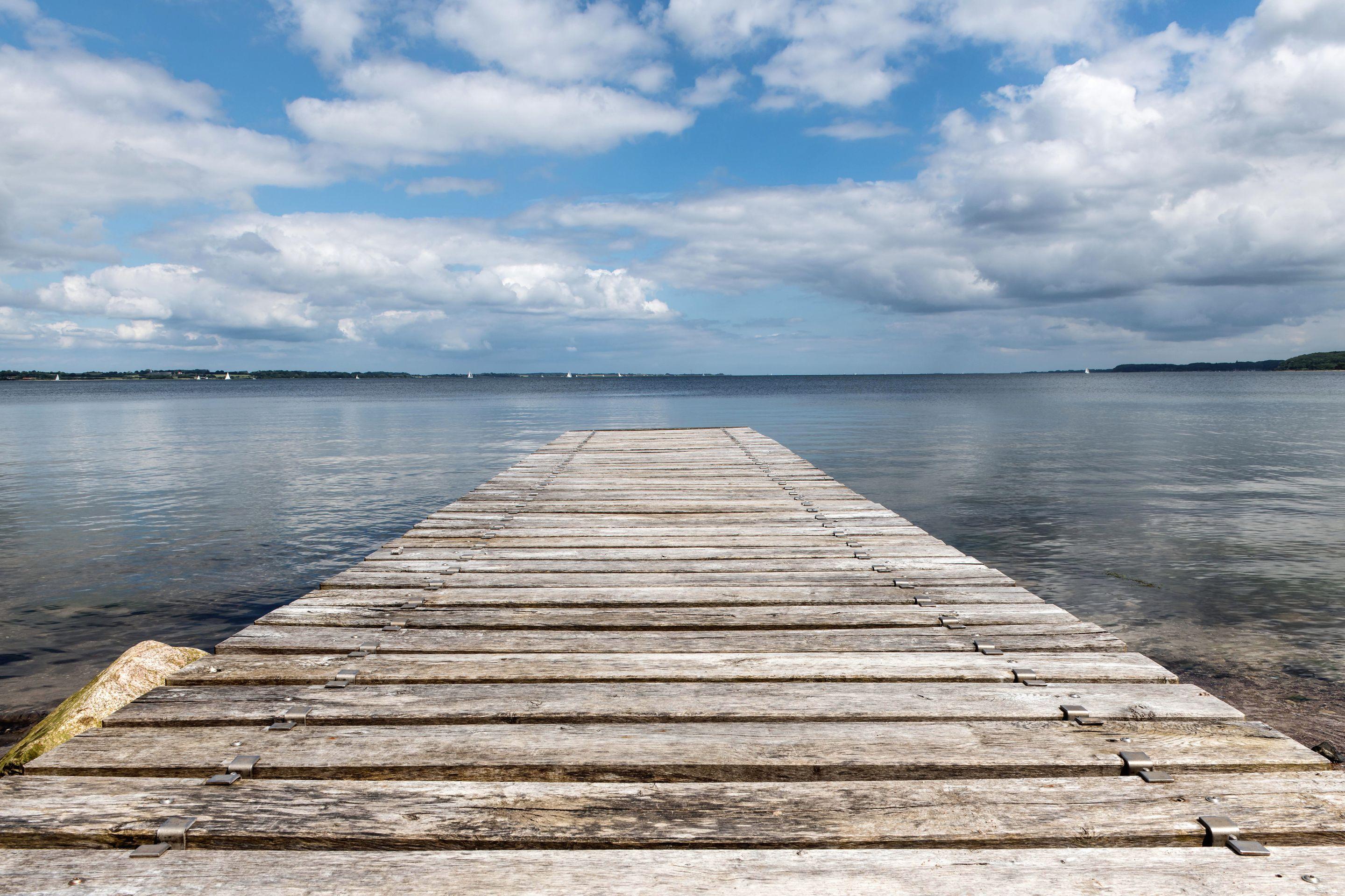 A wooden jetty running out into calm water under a wide sky of white cloud
